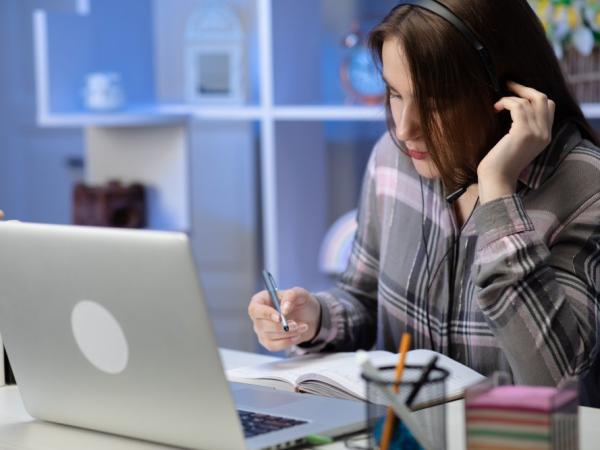 Serious girl student wear headphone study online with internet teacher learn language talk looking at laptop, focused young woman make video call tutoring write notes, teaching concept