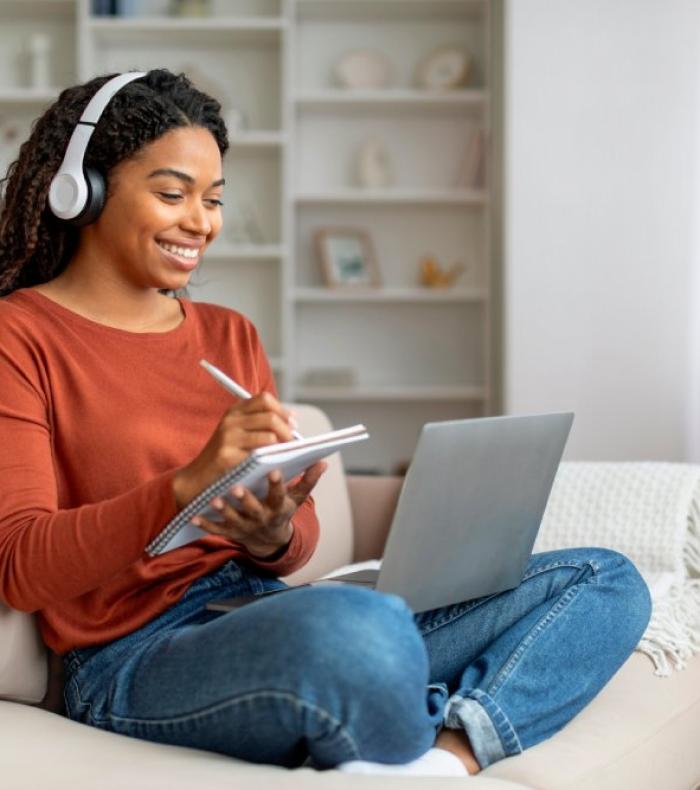 Online Education. Young Black Woman In Headphones Study With Laptop Computer At Home, Smiling African American Female Watching Webinar And Taking Notes To Notepad, Enjoying Distance Learning